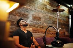 hombre árabe de barba elegante con gafas y camiseta negra fumando narguile en el bar interior. modelo árabe descansando. foto