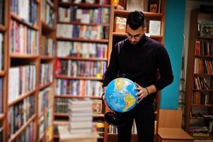 Tall smart arab student man, wear on violet turtleneck and eyeglasses, at library holding earth globe at hands.