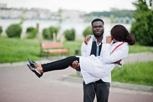 Two young african american doctors couple in white coat. Man hold woman on hands. photo