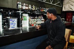 Handsome african american man posing  inside night club in black hat, sitting on bar counter. photo