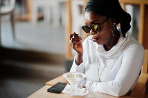 African muslim girl in black hijab and sunglasses sitting at cafe and eating ice cream. photo