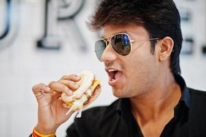 Stylish indian man in sunglasses at fast food cafe eat hamburger against burger sign on wall. photo