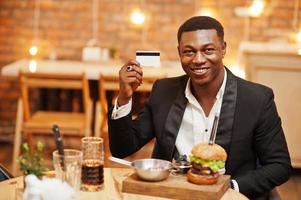 Respectable young african american man in black suit sitting in restaurant hold credit card with tasty double burger and soda drink.