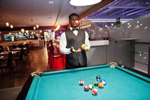 Young handsome african man wearing white shirt, black vest and bow tie play pool billiard. photo