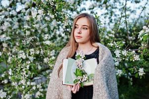 Young brunette girl on plaid against spring blossom tree and holding book at hands. photo