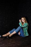 Stylish blonde girl in jacket and jeans against brick black wall at studio. photo