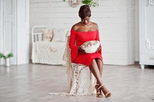 modelo afroamericana de moda en vestido de belleza rojo, mujer sexy posando vestido de noche sentado en una silla en una habitación blanca vintage. foto