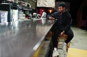 Handsome african american man posing  inside night club in black hat, sitting on bar counter. photo