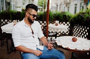 Stylish tall arabian man model in white shirt, jeans and sunglasses posed at street of city. Beard attractive arab guy sitting on outdoor cafe and looking at mobile phone. photo