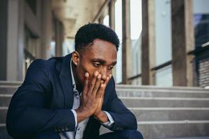 trabajador de oficina masculino cansado después del trabajo, afroamericano tiene dolor de cabeza severo, sentado foto