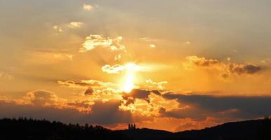 Sunset over the monastery silhouette on the hill. The sun's bright disk is white and backlit clouds. Orange sky and dark purple clouds under. Sky background with place for text. photo