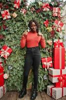 Portrait of a curly haired african woman wearing fashionable red turtleneck posing against christmas decorations, new year eve theme. Show thumbs up. photo