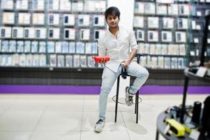 Indian man customer buyer at mobile phone store with wireless earphones and speaker sitting on chair. South asian peoples and technologies concept. Cellphone shop. photo