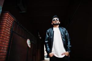 Stylish indian beard man at black leather jacket and sunglasses at tonnel arch. India model posed outdoor at streets of city. photo