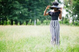 Portrait of gorgeous african american woman 20s in wear in black and white stripe pants and summer hat posing at green grass in park. photo