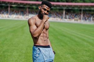 Handsome sexy african american muscular bare torso man at jeans overalls posed at green grass of stadium football field. Fashionable black man portrait. photo