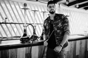 hombre árabe de barba elegante con gafas y chaqueta militar fumando narguile en el bar de la calle. modelo árabe descansando. foto