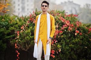 hombre indio con estilo en ropa tradicional amarilla con bufanda blanca, gafas de sol posadas al aire libre contra el árbol de hojas de otoño. foto