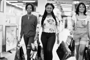 Beautiful three well-dressed afro american girls customers with colored shopping bags walking in tech store. photo