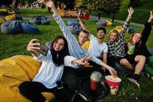 Young multi ethnic group of people watching movie at poof in open air cinema and making selfie on phone. photo