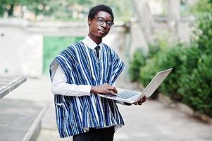 hombre africano con ropa tradicional y gafas con laptop trabajando al aire libre. foto