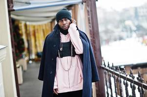Stylish urban style african american man in black coat and hat, pink hoodie posed at winter day. photo