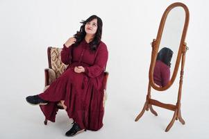 Attractive south asian woman in deep red gown dress posed at studio on white background against mirror and sitting at chair. photo