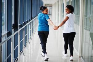 Back of two african woman friends in t-shirts walking halding hands indoor together. photo