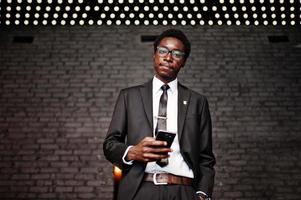 Business african american man wear on black suit and glasses at office looking on mobile phone against futuristic lights. photo