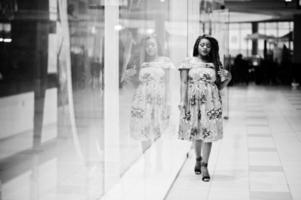 Cute small height african american girl with dreadlocks, wear at coloured yellow dress, posed of shop showcase at trade center. Black and white. photo