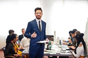 Face of handsome arabian business man, holding clipboard and show thumb up on the background of business peoples multiracial team meeting, sitting in office table. photo
