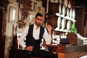 Handsome well-dressed arabian man with glass of whiskey and cigar posed at pub. photo