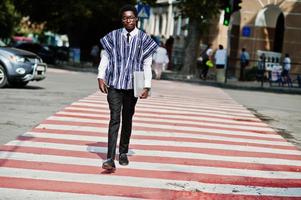 hombre africano con ropa tradicional y anteojos caminando en el cruce de peatones con teléfono móvil y computadora portátil. negocio de África. foto