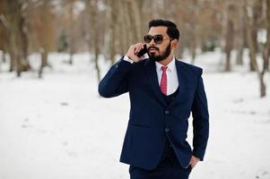 elegante hombre de negocios de barba india con traje y gafas de sol posó en el día de invierno al aire libre, hablando por teléfono móvil. foto