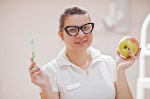 Close up portrait of female dentist woman in glasses standing in her dentistry office with an apple and toothbrush at hands. photo