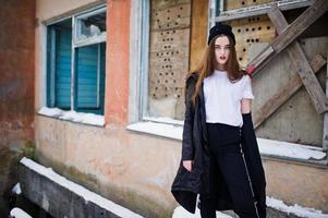 Fashionable long legs brunette model in long black cloak posed outdoor at winter day against old grunge wall with broken windows. photo