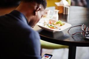 hombre afroamericano de moda en traje sentado en el café y mirando por teléfono. foto