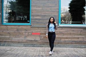 chica afroamericana hipster con camisa de jeans con mangas de leopardo posando en la calle contra una casa de madera con ventanas.