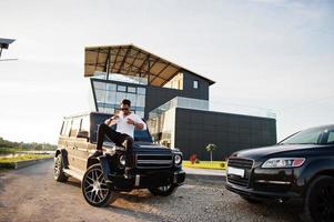 Solid asian man in white shirt and sunglasses posed near black mafia suv car. photo