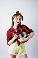 Young funny housewife in checkered shirt and yellow shorts pin up style with saucepan and kitchen spoon isolated on white background. photo