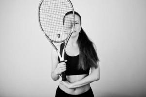 Black and white portrait of beautiful young woman player in sports clothes holding tennis racket while standing against white background. photo