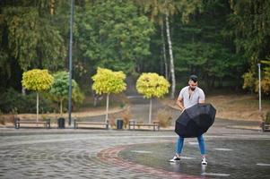 hombre de barba árabe alto de moda usa camisa, jeans y gafas de sol con paraguas posado bajo la lluvia en la plaza del parque. foto