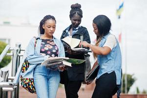 Three african students female posed with backpacks and school items on yard of university and look at book. photo