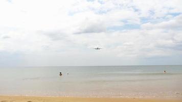 avion de ligne s'approchant de l'aéroport international de phuket au-dessus de l'océan, et des gens le saluant sur la plage en dessous.