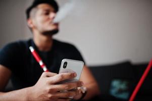 hombre indio guapo y de moda en negro sentado en la habitación, fumando narguile y mirando el teléfono móvil. foto