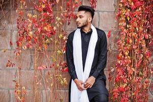 Indian stylish man in black traditional clothes with white scarf posed outdoor against red leaves wall. photo