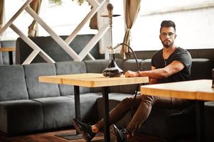 Stylish beard arabian man in glasses and black t-shirt smoking hookah at street bar. Arab model having rest. photo