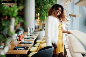 mujer afroamericana glamurosa con vestido amarillo y capa de lana blanca posada en el restaurante cerca de las ventanas. foto