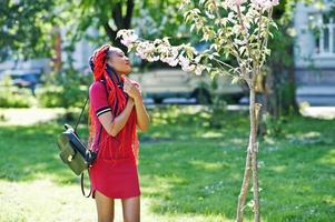 Cute and slim african american girl in red dress with dreadlocks posed outdoor in spring park. Stylish black model. photo