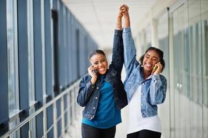 Two african woman friends in jeans jacket speak on phone and hands up indoor together. photo
